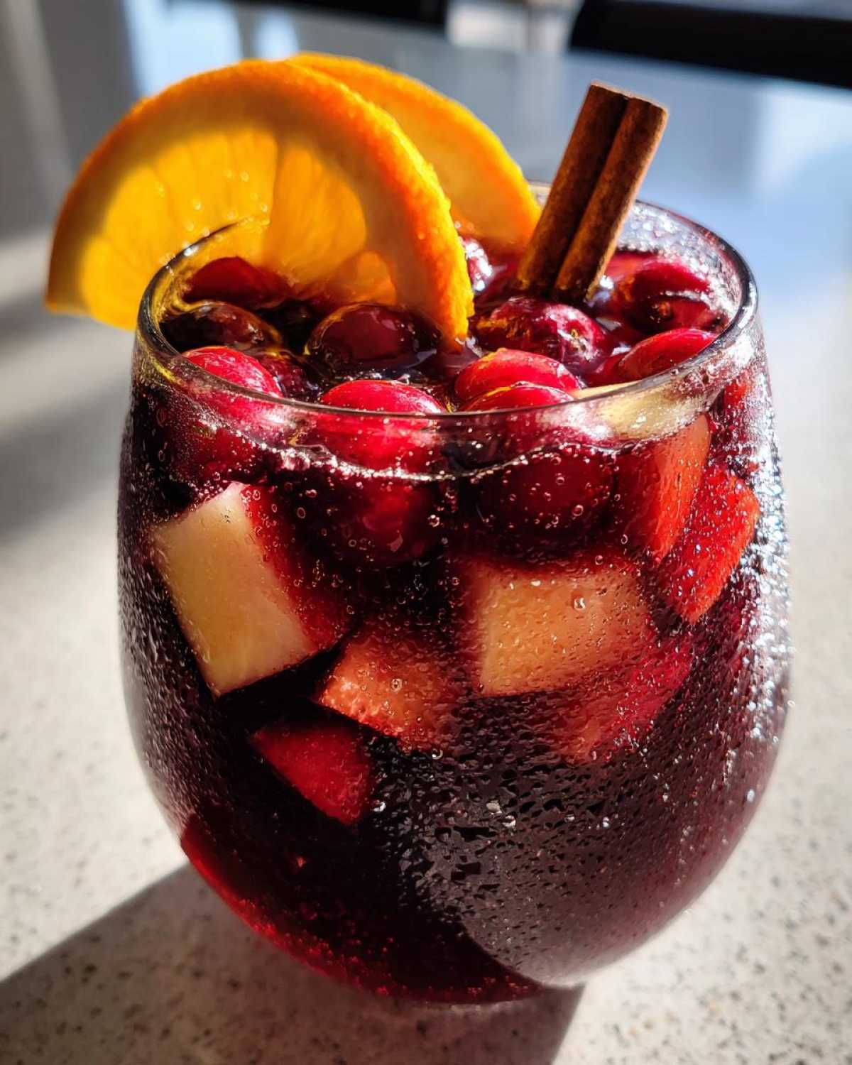 A close-up of a chilled glass of festive christmas sangria, garnished with orange slices, cranberries, and a cinnamon stick.