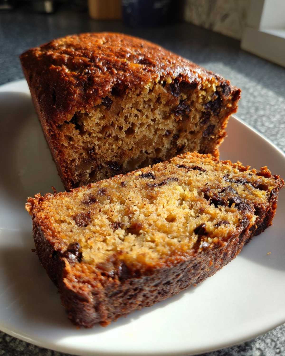 A moist slice of chocolate chip vegan banana bread next to the main loaf on a white plate, showing a golden crumb.