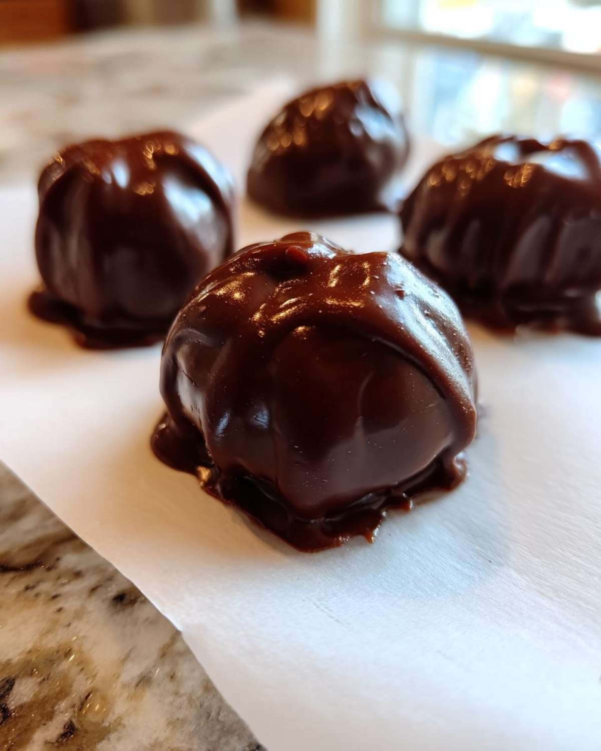 Close-up of four glossy, dark chocolate covered cherries resting on white parchment paper.