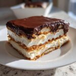 A square slice of layered chocolate eclair cake featuring graham crackers, white cream filling, and a thick chocolate glaze.