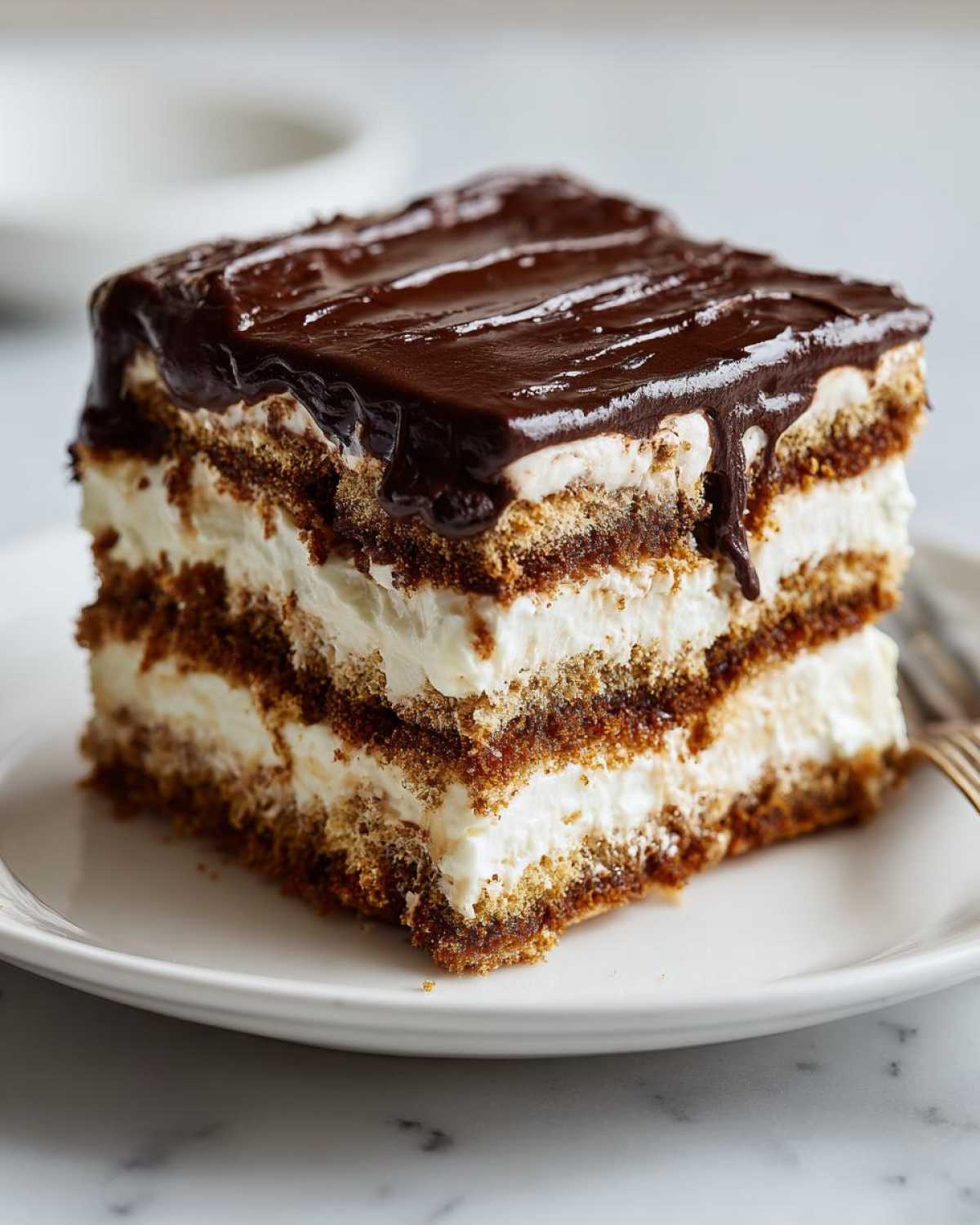 A square slice of layered chocolate eclair cake with graham cracker layers and creamy filling, topped with rich chocolate glaze.