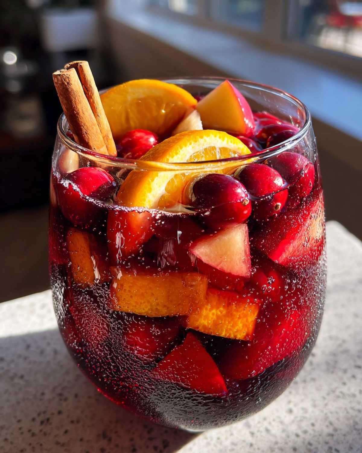 A glass filled with vibrant red christmas sangria, loaded with cranberries, orange slices, apples, and two cinnamon sticks.