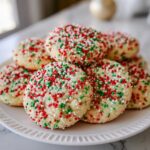A stack of freshly baked christmas sprinkle sugar cookies covered in red, green, and white nonpareils on a white plate.