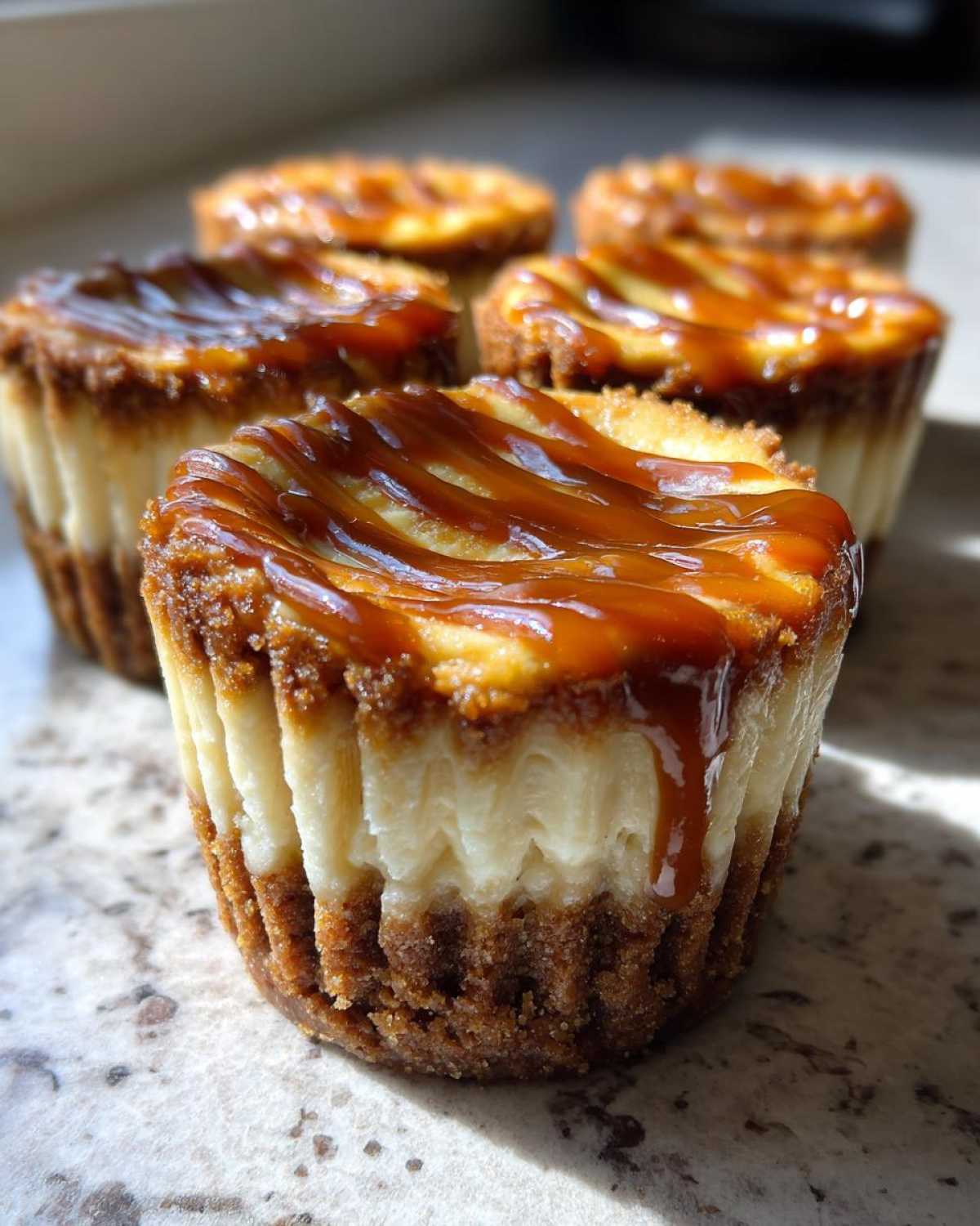 A close-up of one of the cookie butter cheesecake cups topped with a rich caramel drizzle, with more cups blurred in the background.
