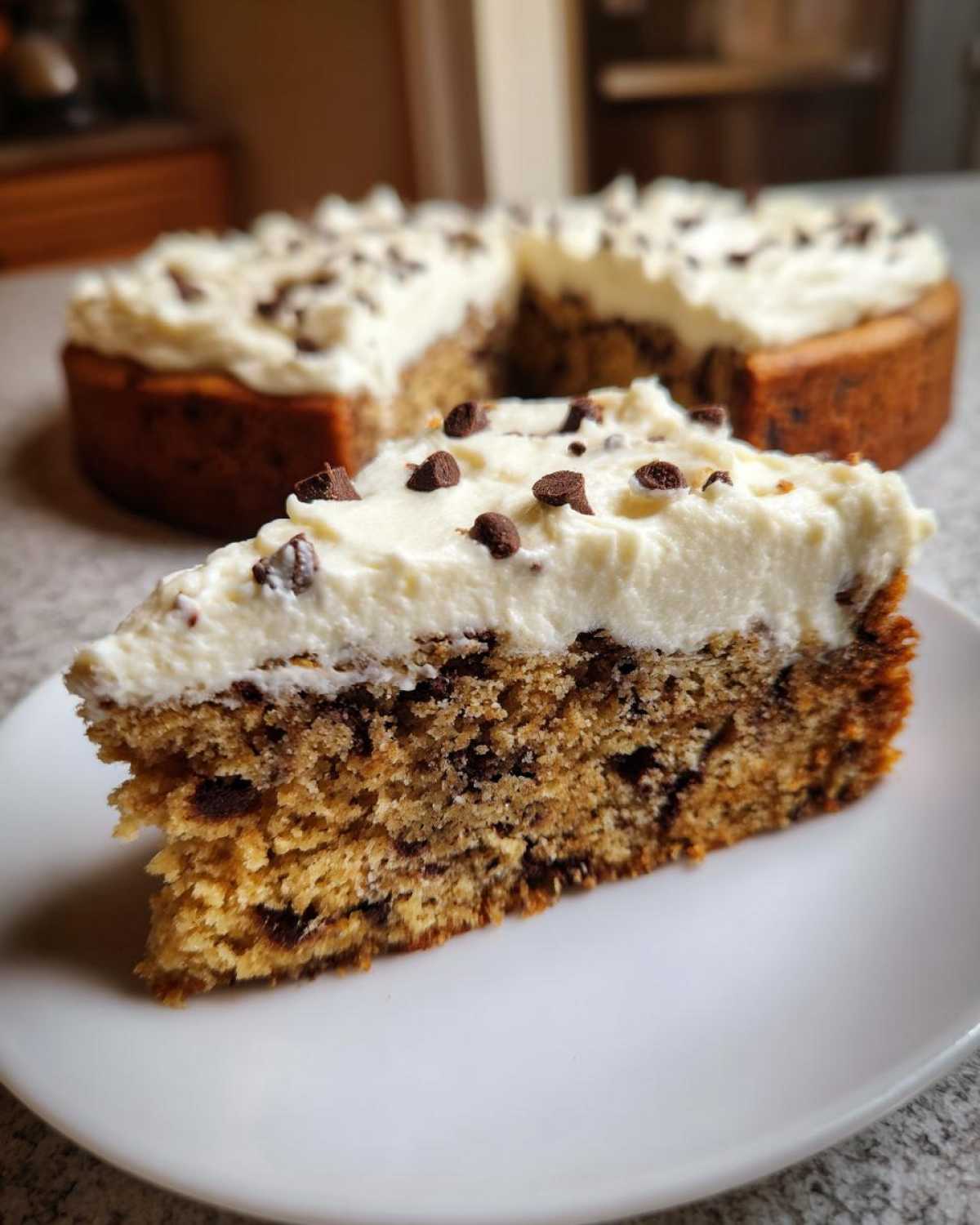 A thick slice of chocolate chip cookie cake recipe topped with white frosting and mini chocolate chips.