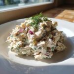 A mound of creamy cottage cheese chicken salad mixed with red onion and topped with fresh dill on a white plate.