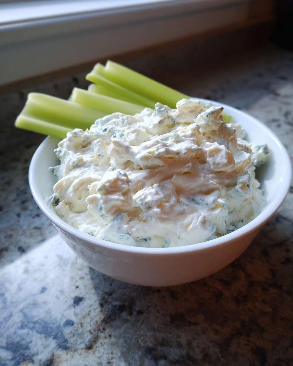 A white bowl filled with chunky, creamy blue cheese dip, served alongside fresh celery sticks.