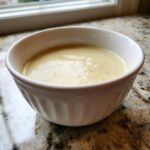 A close-up of a white, fluted bowl filled with smooth, creamy cauliflower soup, set on a granite countertop.