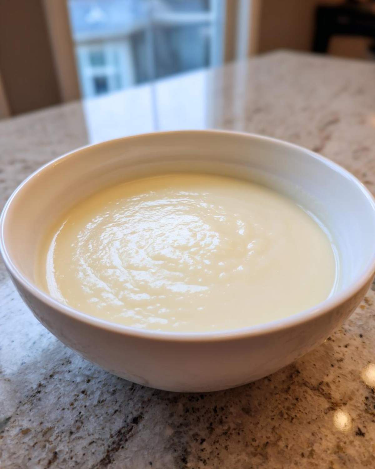 Close-up of a white bowl filled with smooth, creamy cauliflower soup resting on a granite countertop.