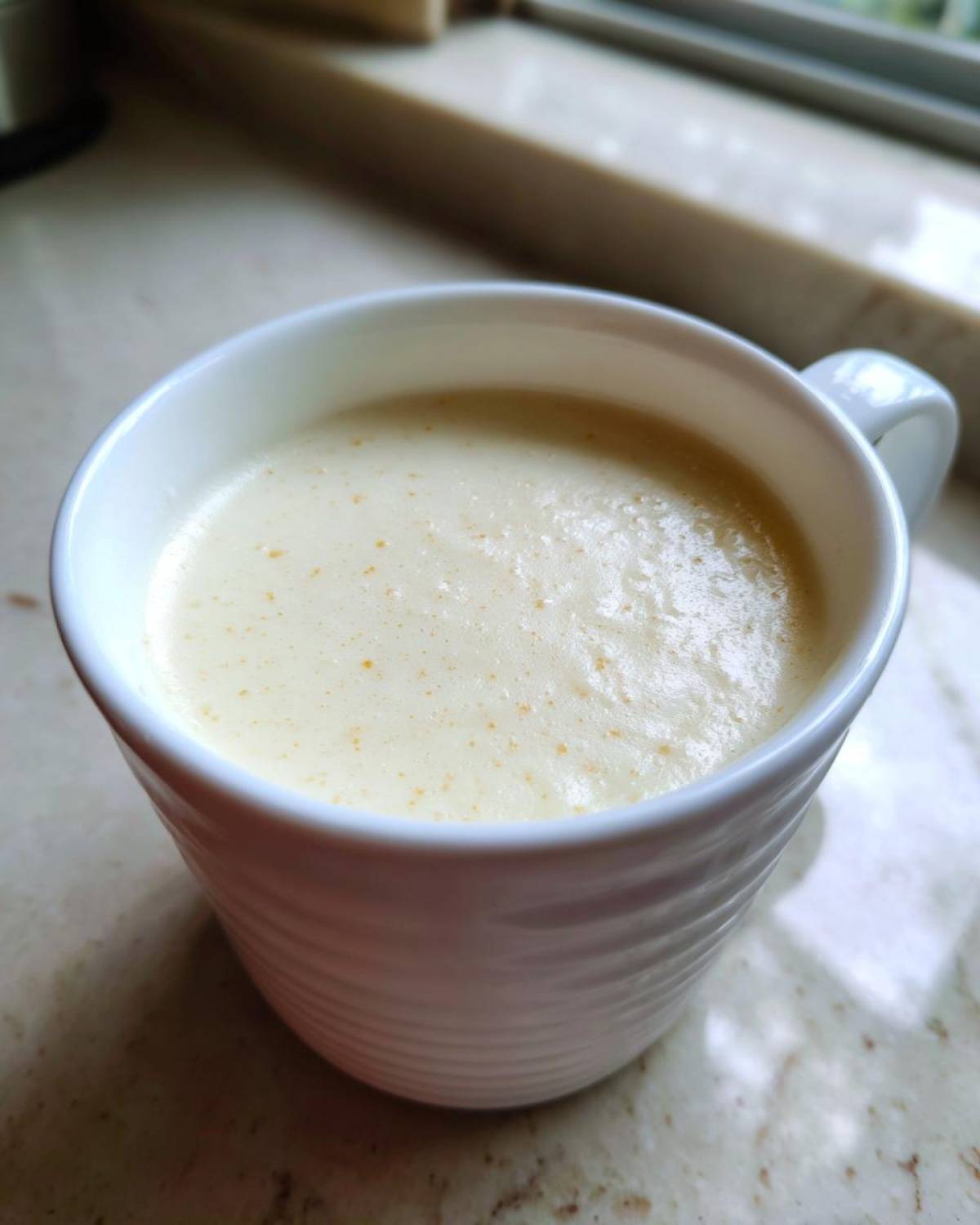 Close-up of creamy, light yellow cauliflower soup served hot in a white, textured mug.