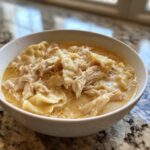 Close-up of a white bowl filled with creamy chicken and dumplings, featuring shredded chicken and thick noodles.