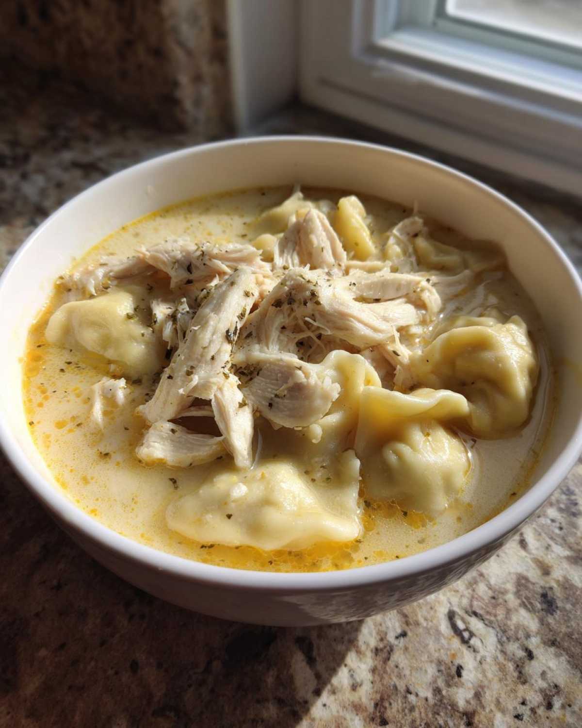A close-up of a white bowl filled with creamy chicken and dumplings, topped with shredded chicken and herbs.