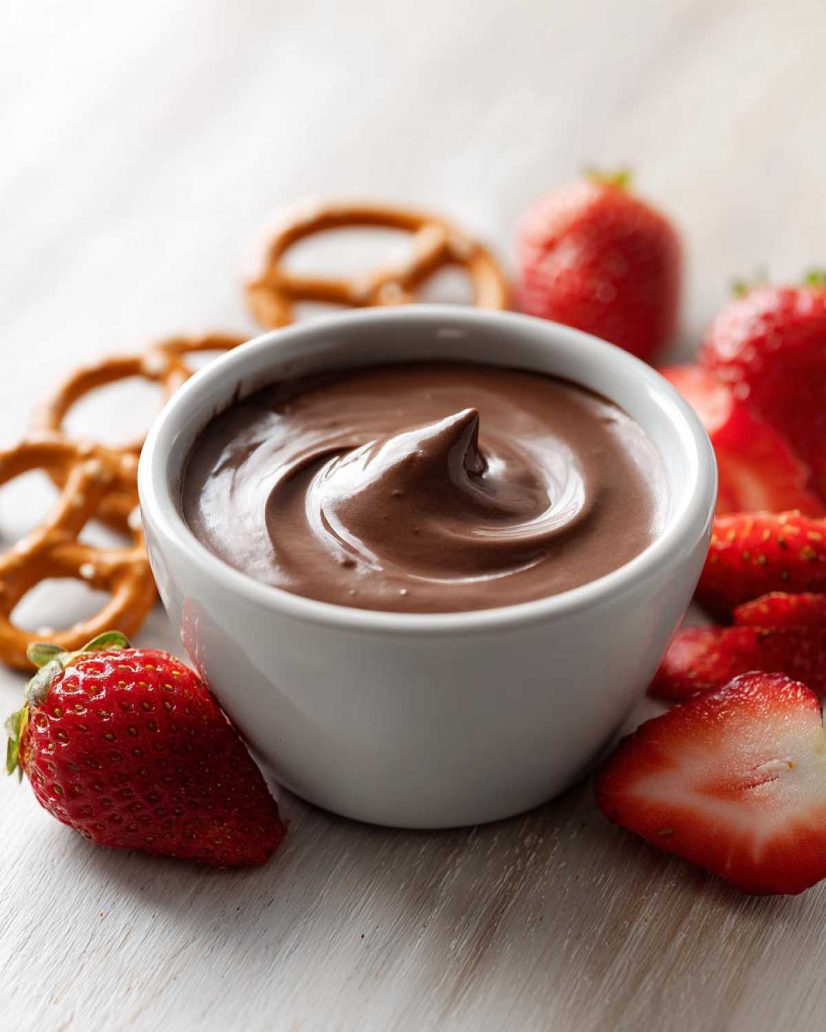 A small white bowl filled with smooth, rich chocolate dip, surrounded by fresh strawberries and pretzels.