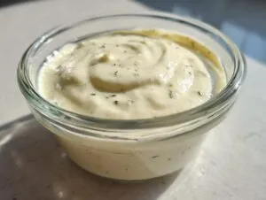 Close-up of creamy, pale yellow horseradish sauce with flecks of green herbs in a small glass bowl.