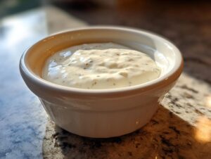 A close-up of thick, white creamy horseradish sauce speckled with herbs, served in a small white dipping bowl.
