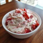 A close-up of creamy strawberry cheesecake dip topped with fresh strawberry chunks in a white bowl.