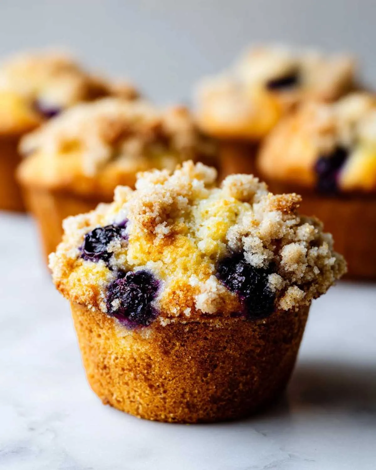 A close-up of a golden brown blueberry muffin topped with sweet streusel crumbs, with more blueberry muffins blurred in the background.