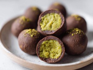 Close-up of several rich, chocolate-coated dubai chocolate balls, one cut in half revealing a bright green pistachio filling.