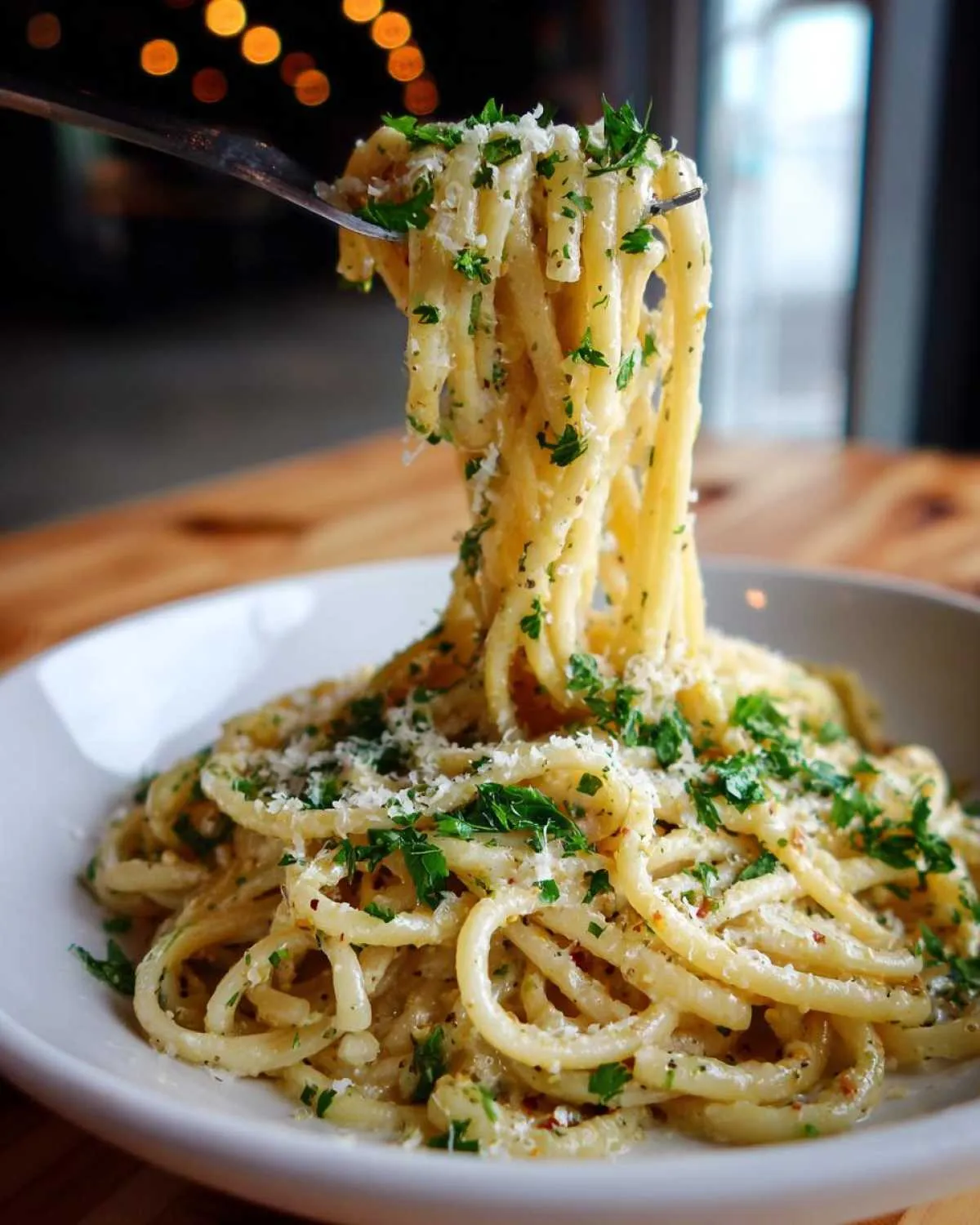 A fork lifts long strands of creamy garlic parmesan pasta topped with grated cheese and fresh parsley from a white bowl.
