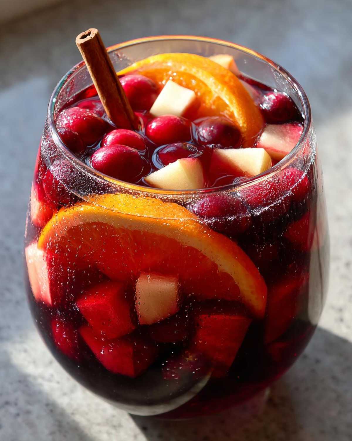 Close-up of a glass filled with rich red christmas sangria, garnished with orange slices, cranberries, apple chunks, and a cinnamon stick.