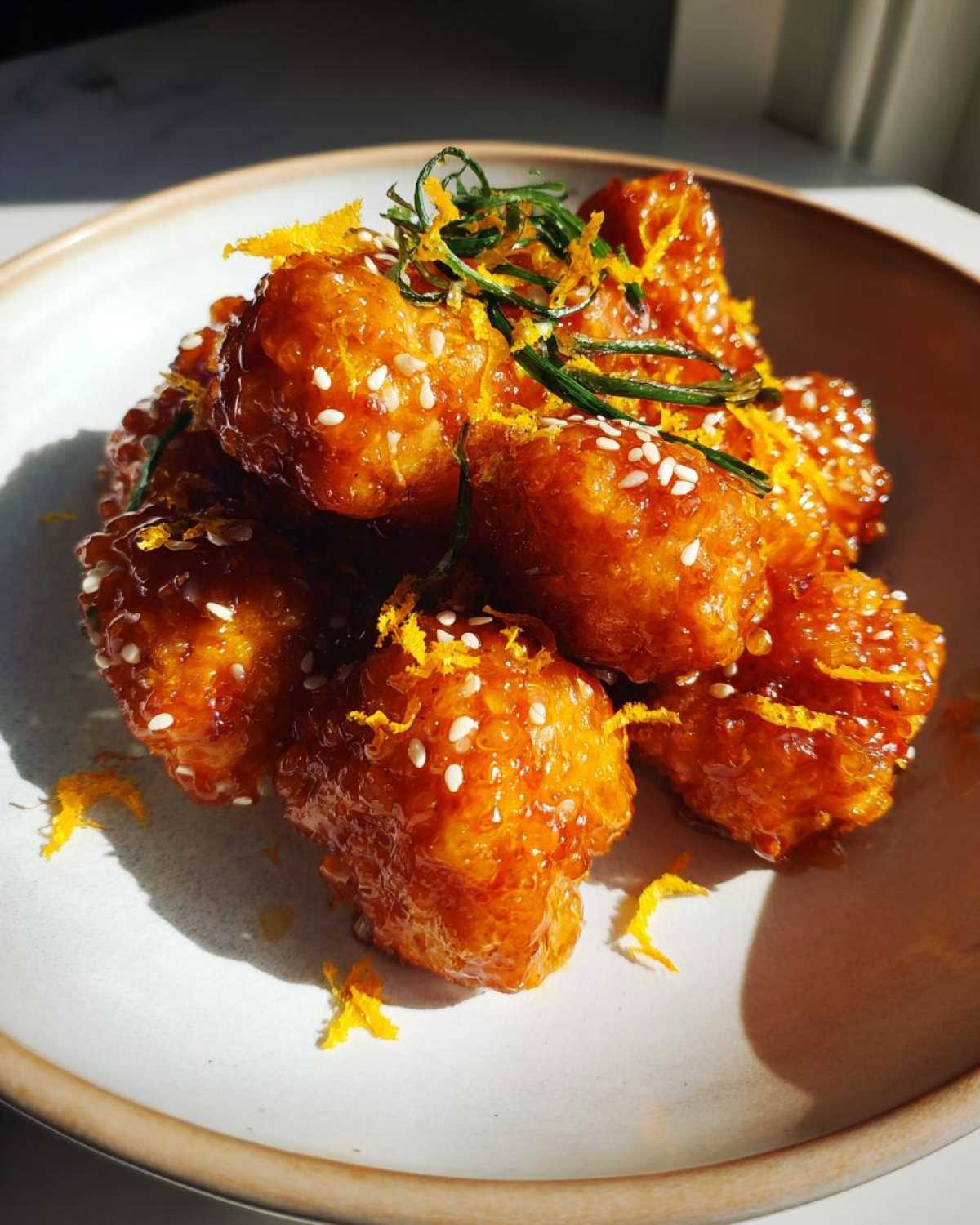 Close-up of sticky, glazed orange chicken recipe pieces topped with sesame seeds and orange zest.