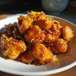 Close-up of crispy, saucy orange chicken recipe pieces garnished with sesame seeds and bright orange zest.