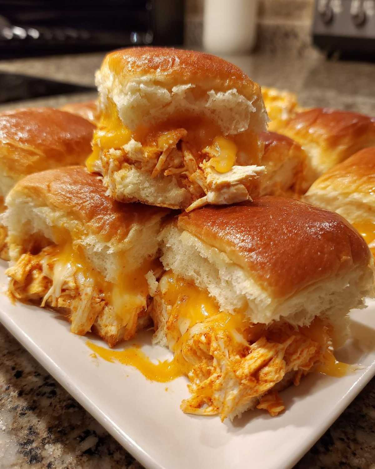 A close-up of several gooey buffalo chicken sliders stacked on a white plate, showing melted cheddar cheese.