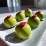 Six shiny, bright green Grinch Oreo Balls topped with small red hearts, arranged on a white platter.