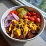 Close-up of a high-protein cheeseburger bowl featuring seasoned ground beef, melted cheese sauce, red onion, pickles, and cherry tomatoes.