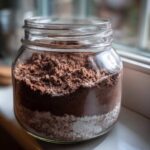 A clear glass jar filled with layered homemade hot cocoa mix, showing dark brown cocoa powder over white sugar.