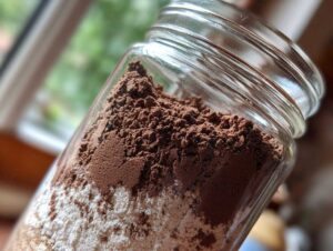 Close-up of layered dry ingredients, including cocoa powder and white powder, for homemade hot cocoa mix in a clear glass jar.