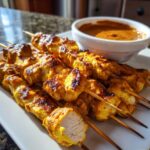 Close-up of grilled, golden chicken satay skewers served with a side of rich peanut dipping sauce.