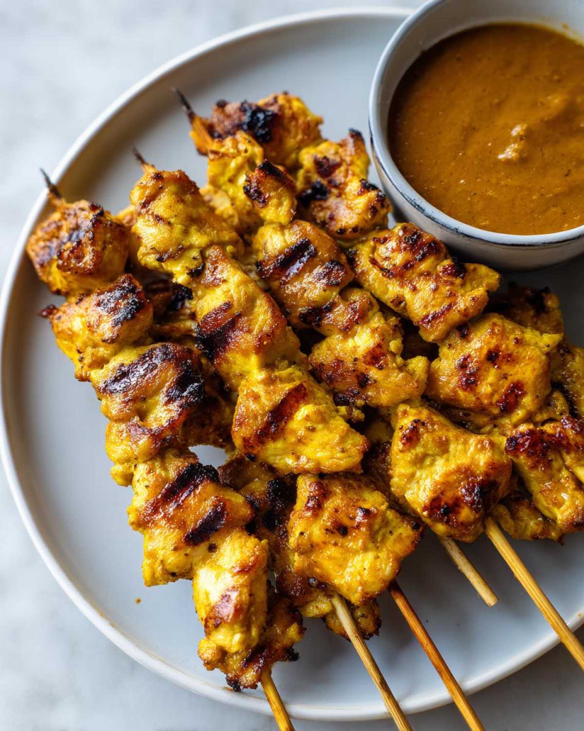 Close-up of grilled, yellow-marinated chicken satay skewers served on a plate next to a bowl of peanut sauce.
