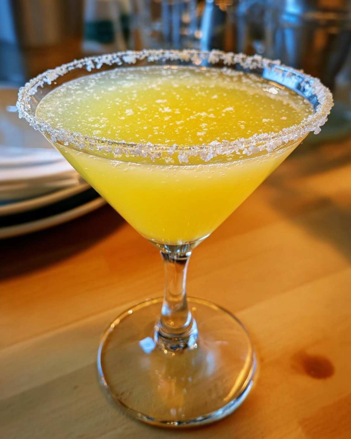 Close-up of a bright yellow lemon drop martini in a coupe glass with a sugar-rimmed edge, sitting on a wooden table.