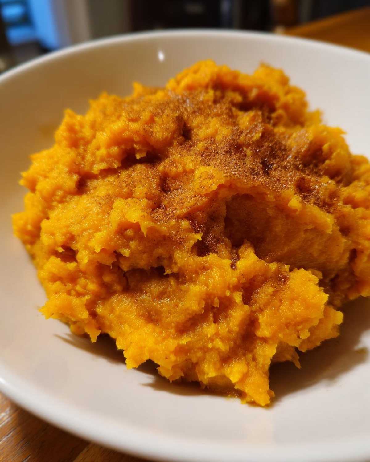 Close-up of fluffy, bright orange mashed sweet potatoes topped with a sprinkle of cinnamon in a white bowl.