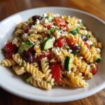 Close-up of a bowl filled with mediterranean pasta salad featuring rotini, cherry tomatoes, cucumber, olives, and feta cheese.