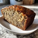 A close-up of a thick slice of moist vegan banana bread with visible dark specks, likely chocolate chips, on a white plate.