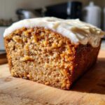 Close-up of a thick slice of moist spice cake topped with creamy white frosting, resting on a wooden board.