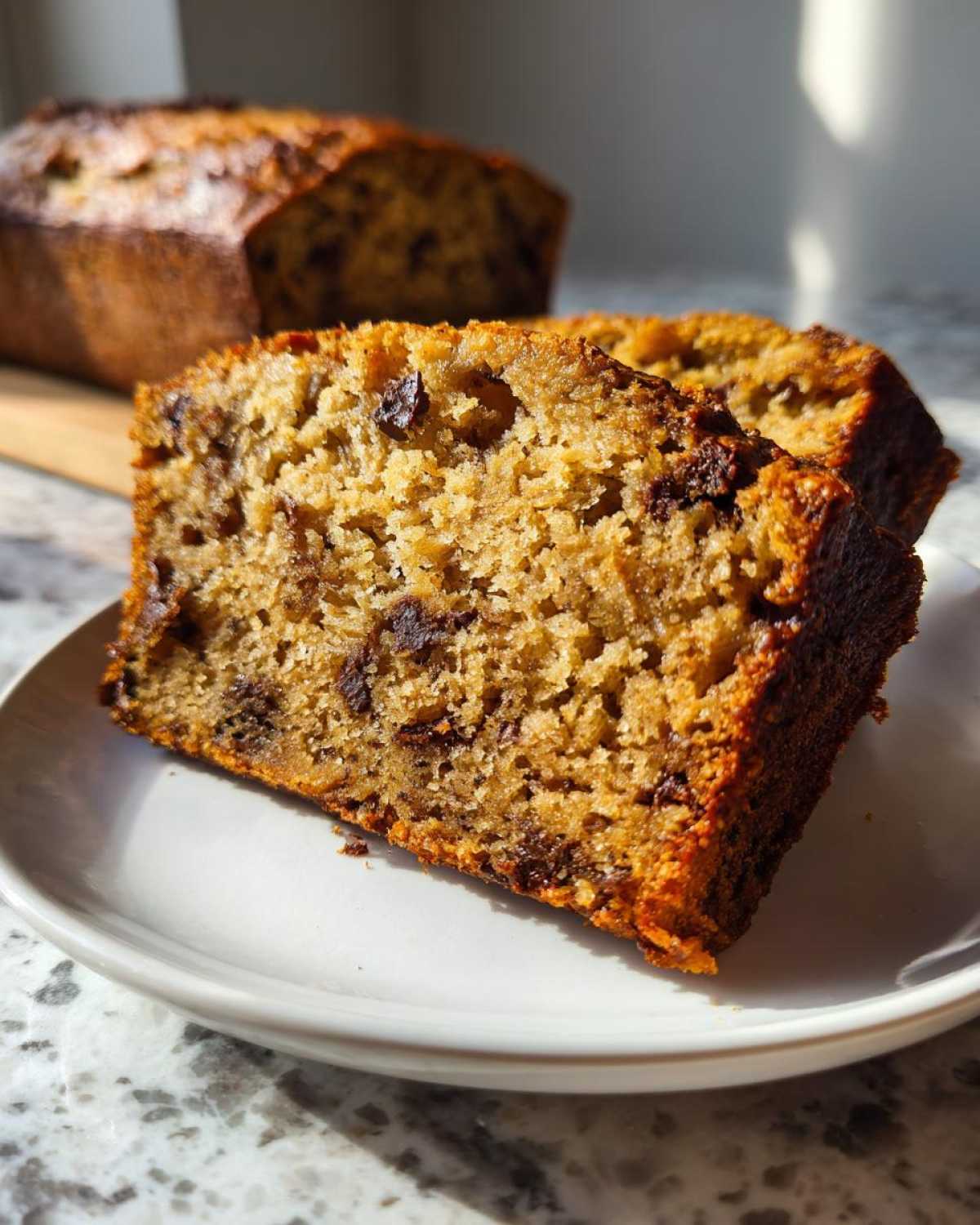 A close-up of two slices of moist vegan banana bread with visible chocolate chips, served on a white plate.