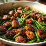 Close-up of glazed chicken pieces mixed with bright green beans in a skillet, featuring a rich, dark sauce.