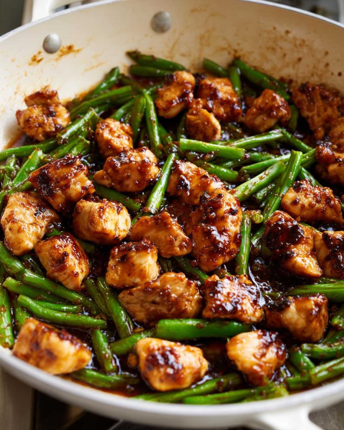 Close-up of bite-sized chicken pieces coated in a dark glaze mixed with bright green beans in a white skillet, showing the finished chicken and green beans dish.