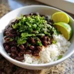 A white bowl filled with fluffy white rice topped generously with seasoned black beans and fresh cilantro, served with lime wedges for the perfect beans and rice.