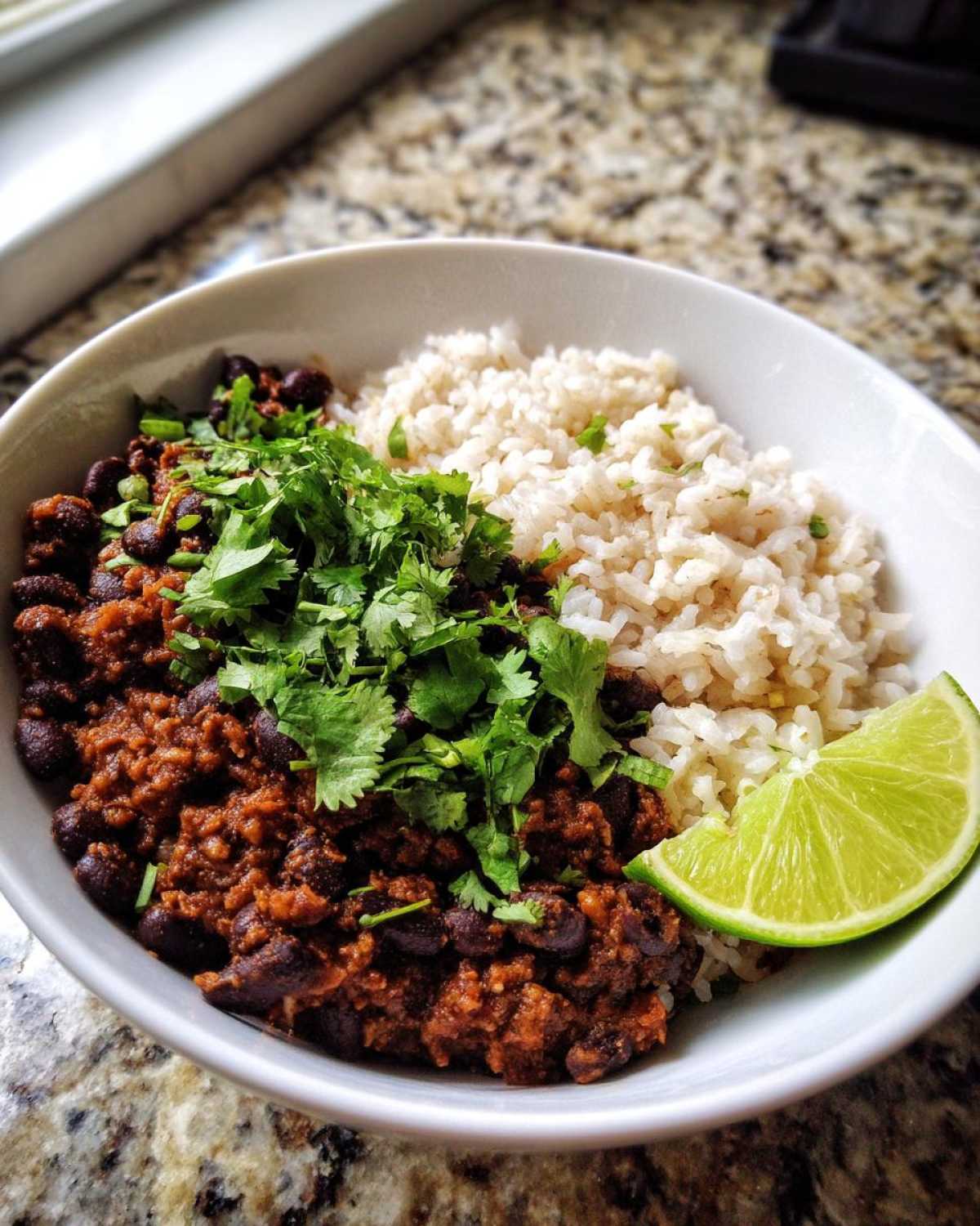 A white bowl filled with seasoned black beans and rice, topped generously with fresh cilantro and a lime wedge, showcasing a perfect beans and rice meal.