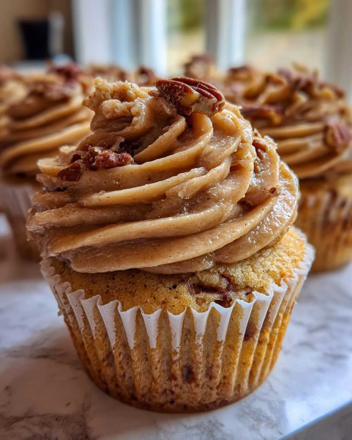 A close-up of a freshly baked pecan pie cupcake topped with thick, swirled brown sugar frosting and chopped pecans.