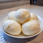 A white plate holding several smooth, pale yellow mounds of freshly made fufu.