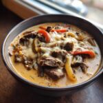 Close-up of a hearty bowl of creamy philly cheese steak soup topped with melted white cheese, steak pieces, and bell peppers.