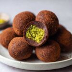 Close-up of several dubai chocolate balls dusted with cocoa powder, one cut open revealing a bright green pistachio filling.