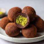 Close-up of several dubai chocolate balls dusted with cocoa powder, one cut open revealing a bright green pistachio filling.