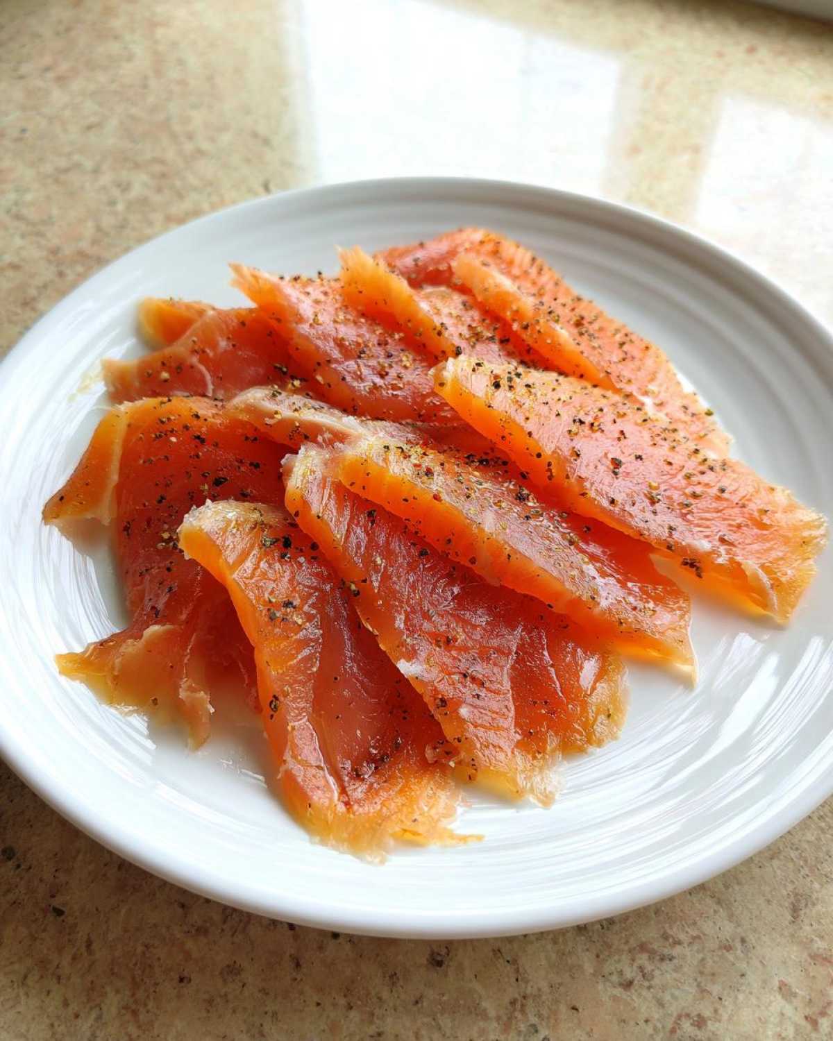 Close-up of thinly sliced smoked salmon seasoned with cracked black pepper on a white plate.