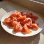 A white plate piled high with thinly sliced, bright orange smoked salmon seasoned with pepper.